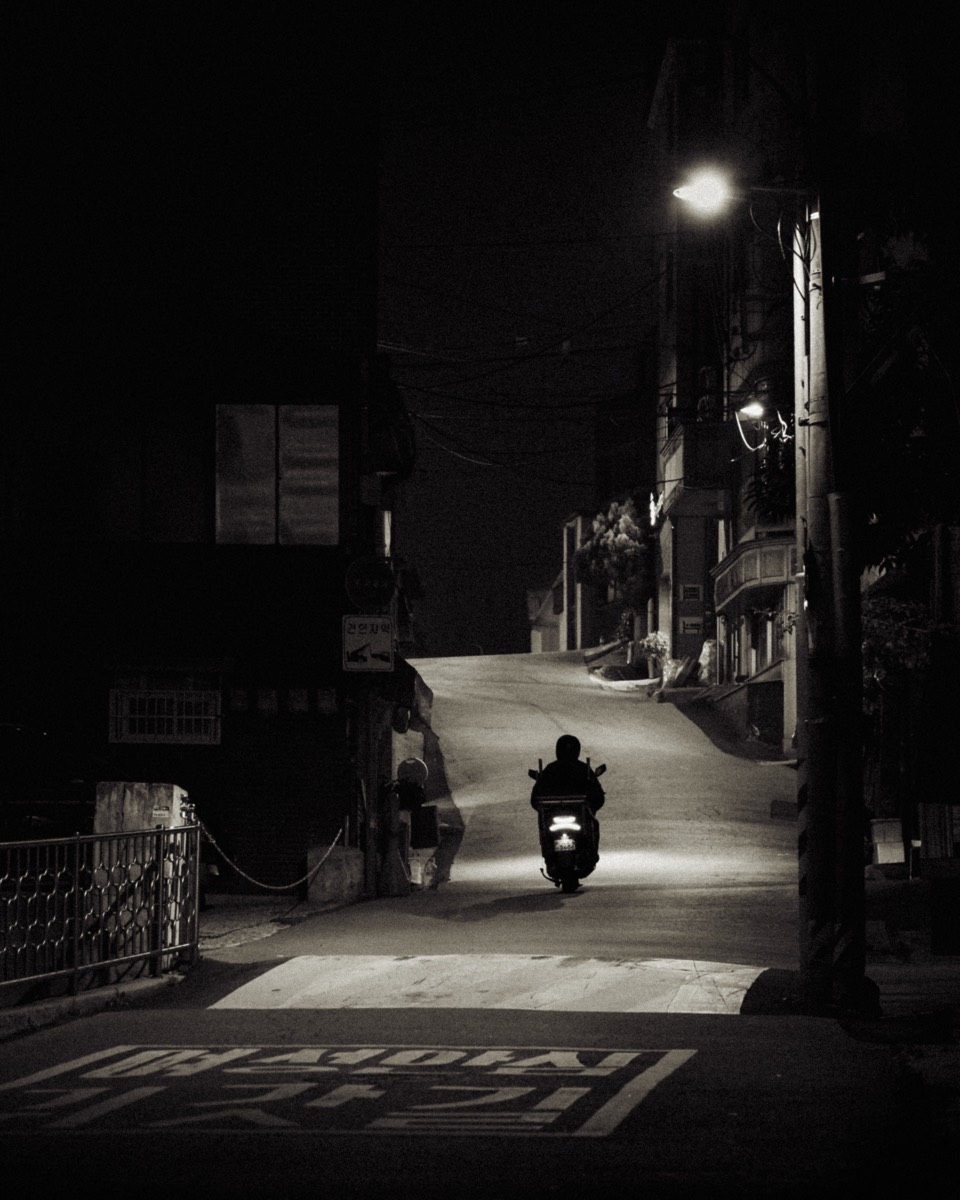 A lone scooter rider rolls down a quiet sloped alley under a single streetlight at night.