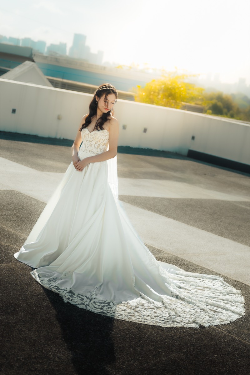 A bride in a long gown stands on a rooftop, her train fanned out, the city skyline behind her in warm backlight.