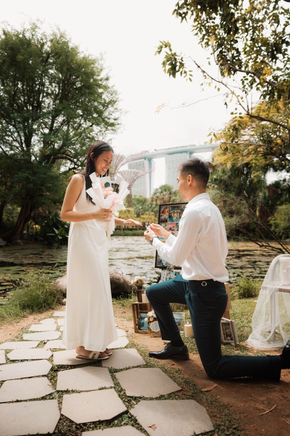 A man kneels to propose on a garden path by the water, his partner holding a bouquet, the city beyond.