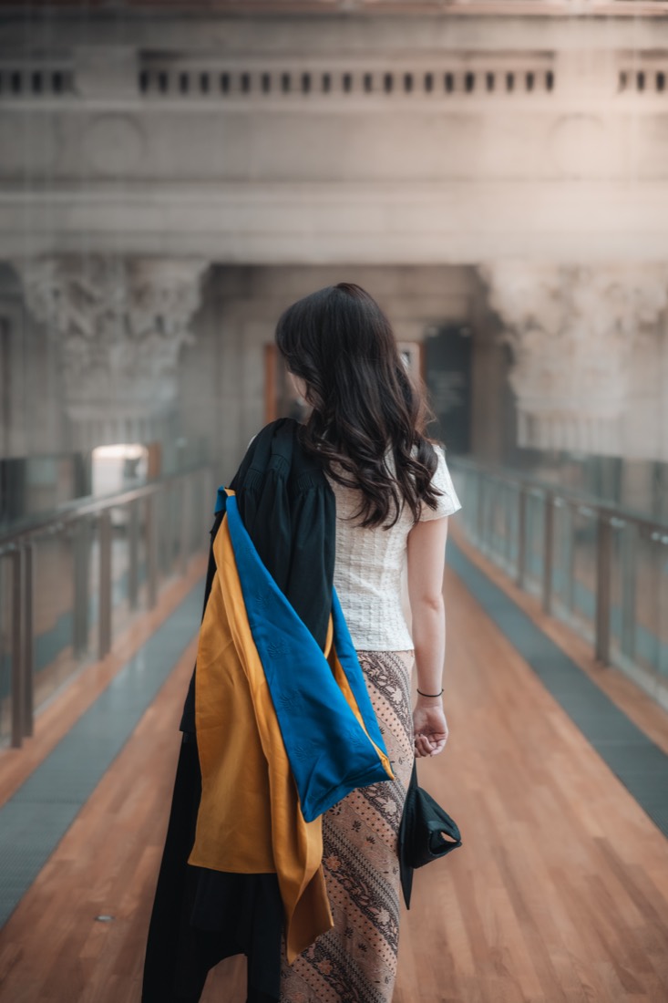 A graduate seen from behind, her gown slung over one shoulder, walking through a grand pillared hall.