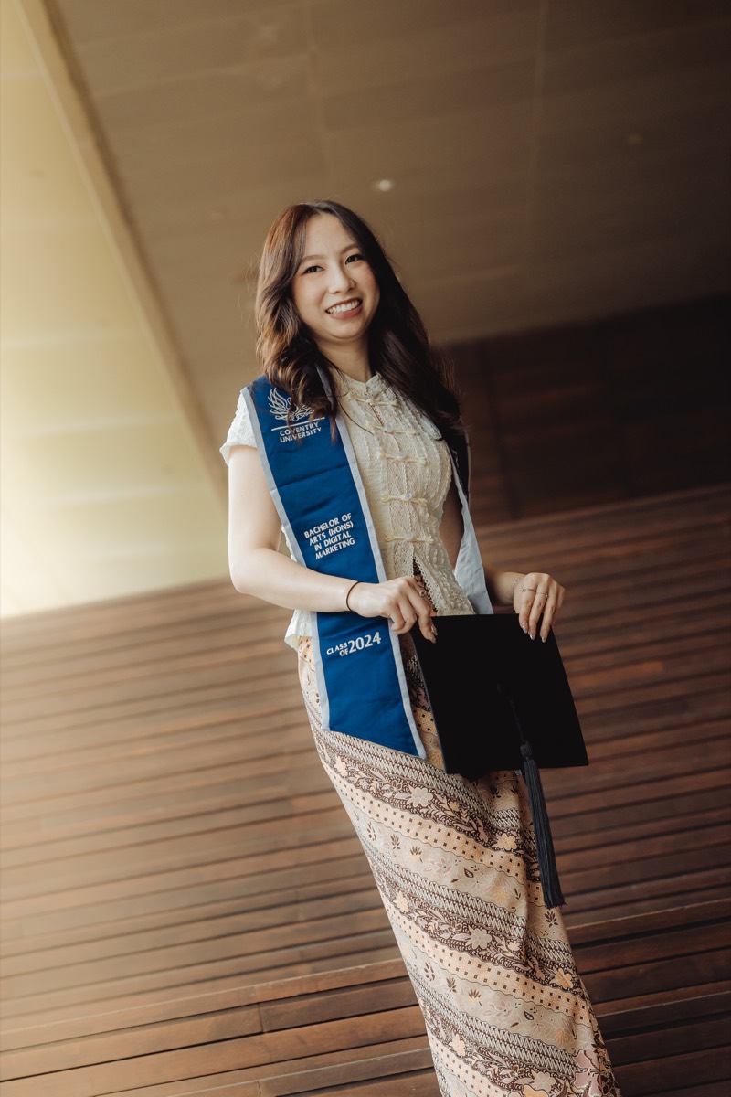 A graduate stands full-length on a boardwalk, holding her cap.