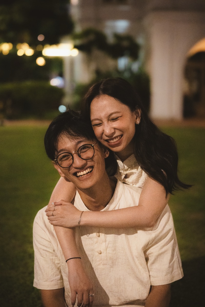A couple embrace from behind on a lawn at night, both laughing.