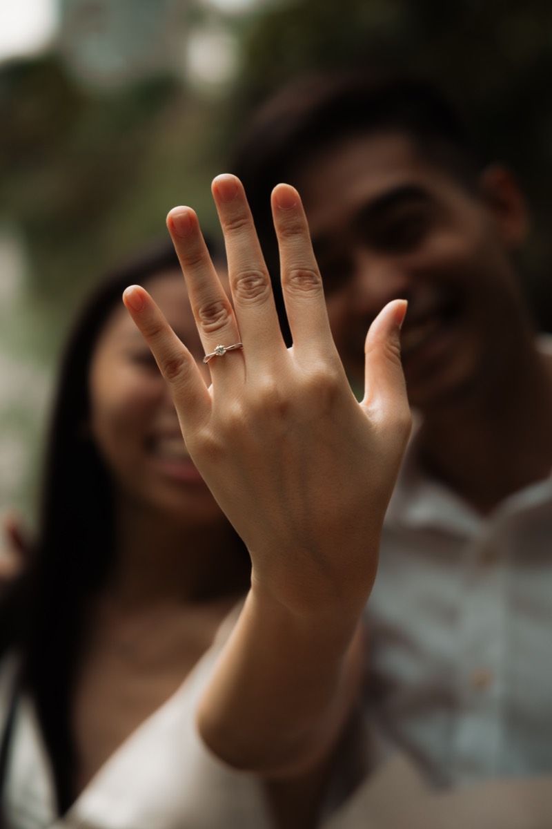 Her hand and new ring held up to the camera, the couple laughing out of focus behind.