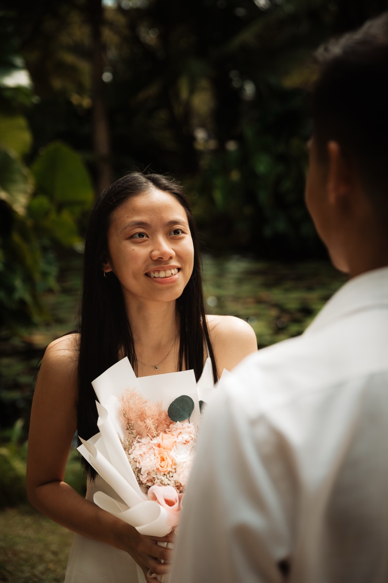 A woman holds a bouquet and meets her partner's eyes in a green garden.