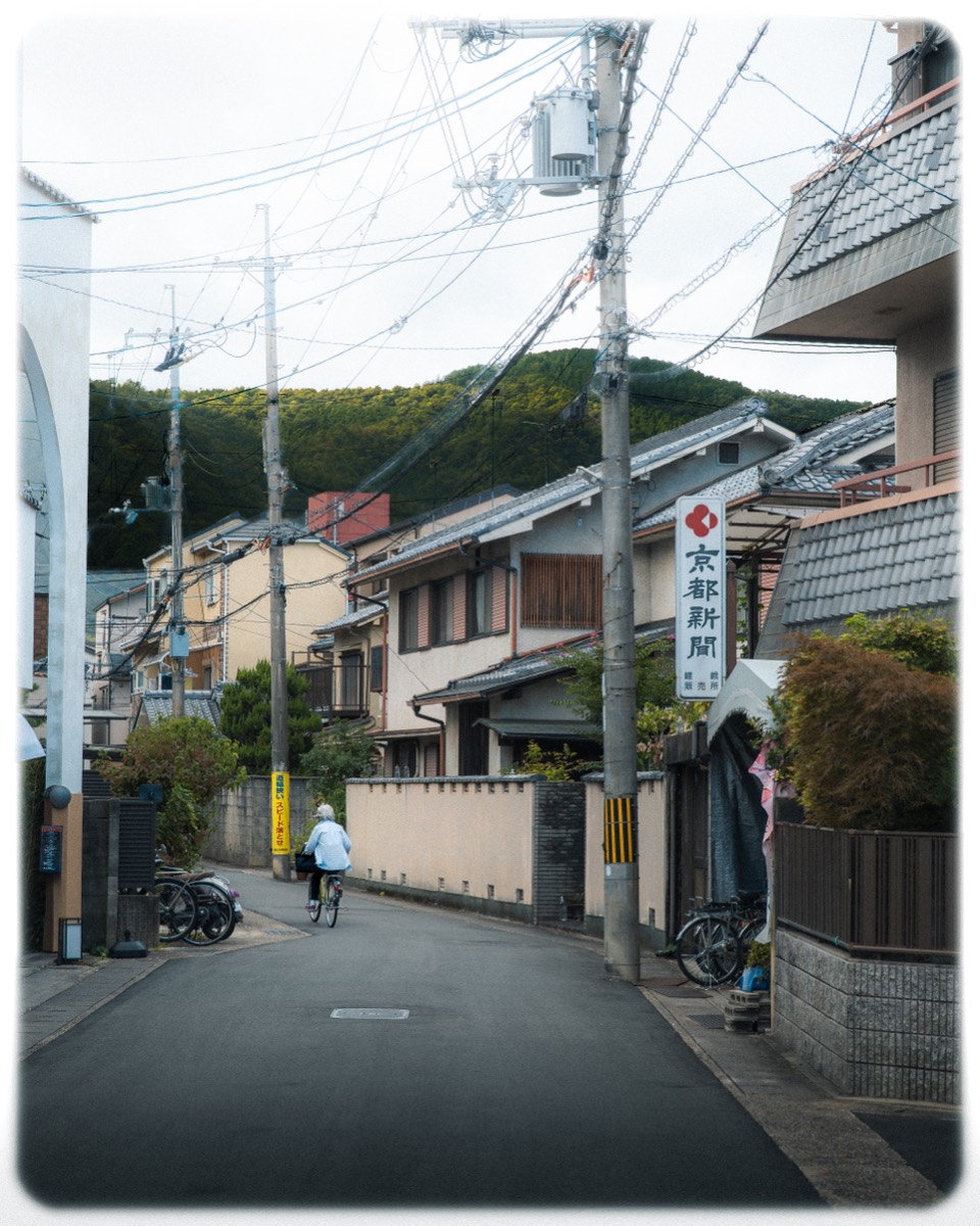 A cyclist rides a quiet residential lane beneath a wooded hill.