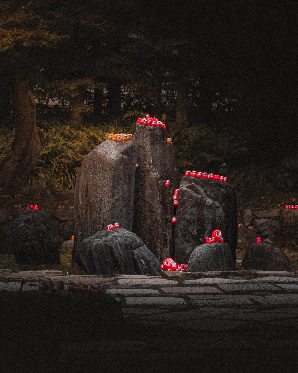 Red offerings and candles glow among dark stones at a forest shrine.