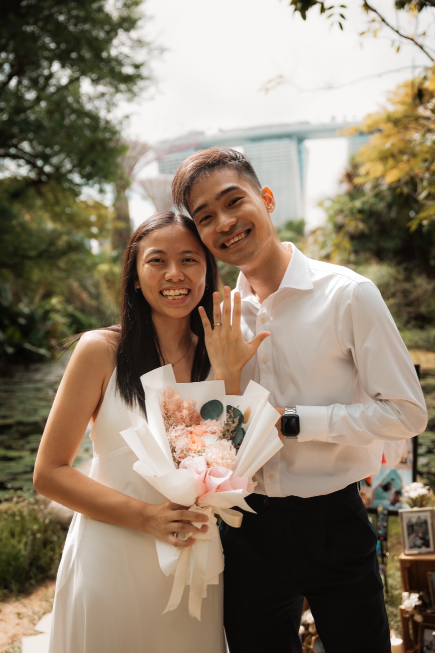 A newly engaged couple smile together, the woman holding a bouquet, the city behind them.