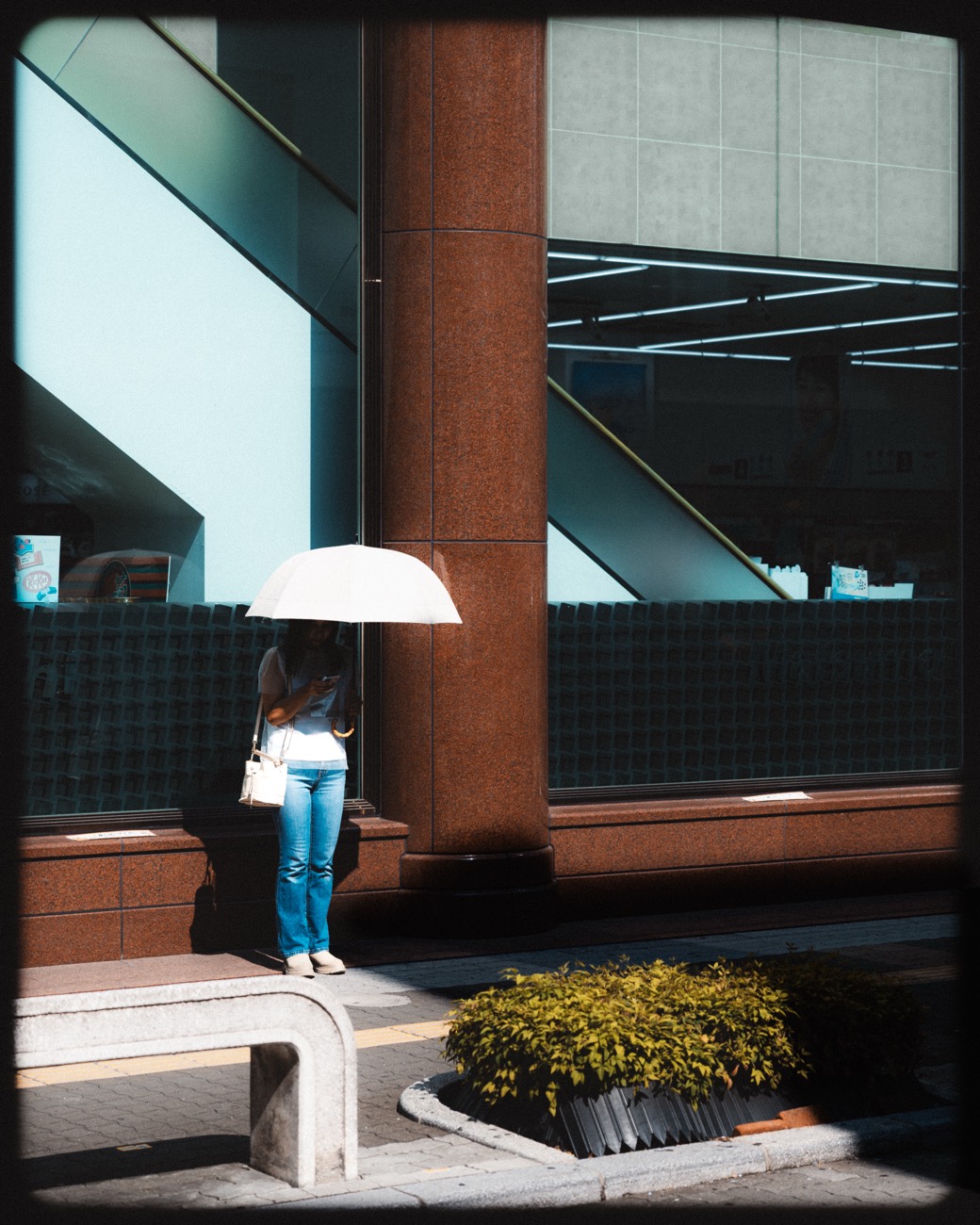 A person under a white umbrella waits beside a column, caught in teal and red afternoon shadow.