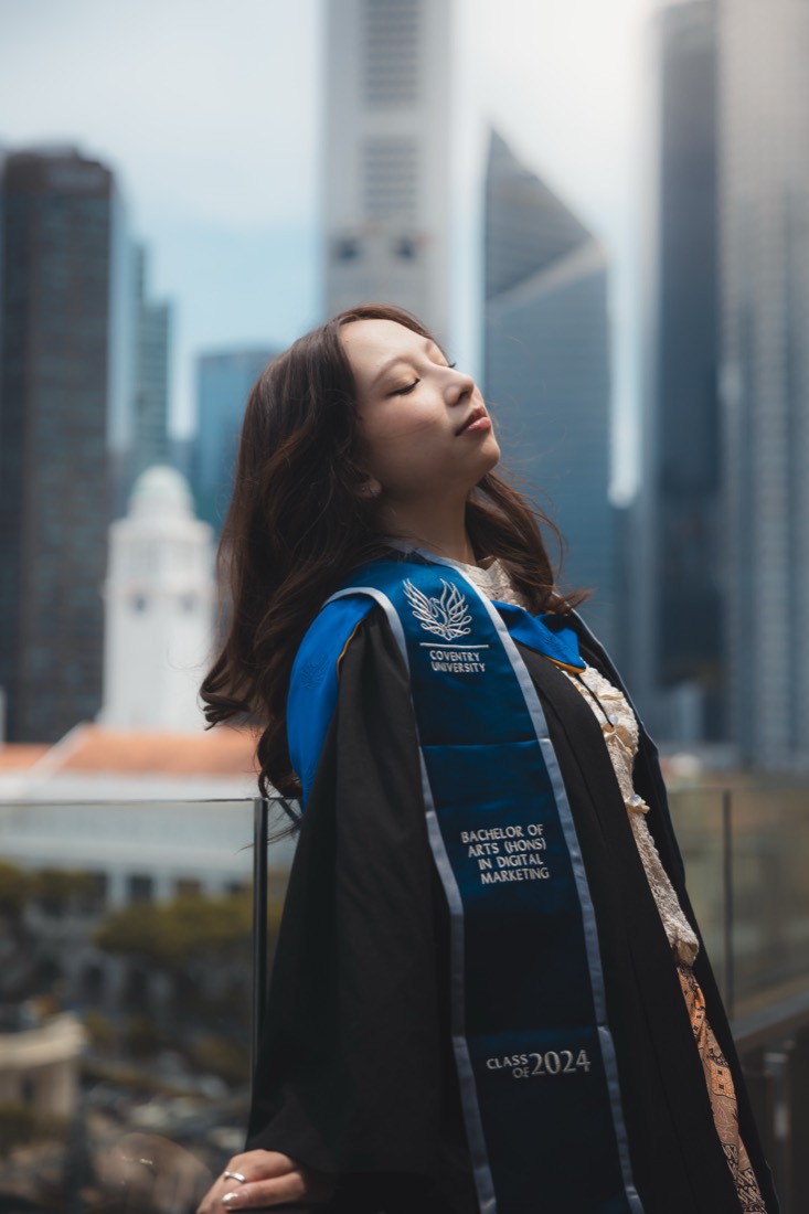 A graduate in cap and gown tilts her head back, eyes closed, the city skyline soft behind her.