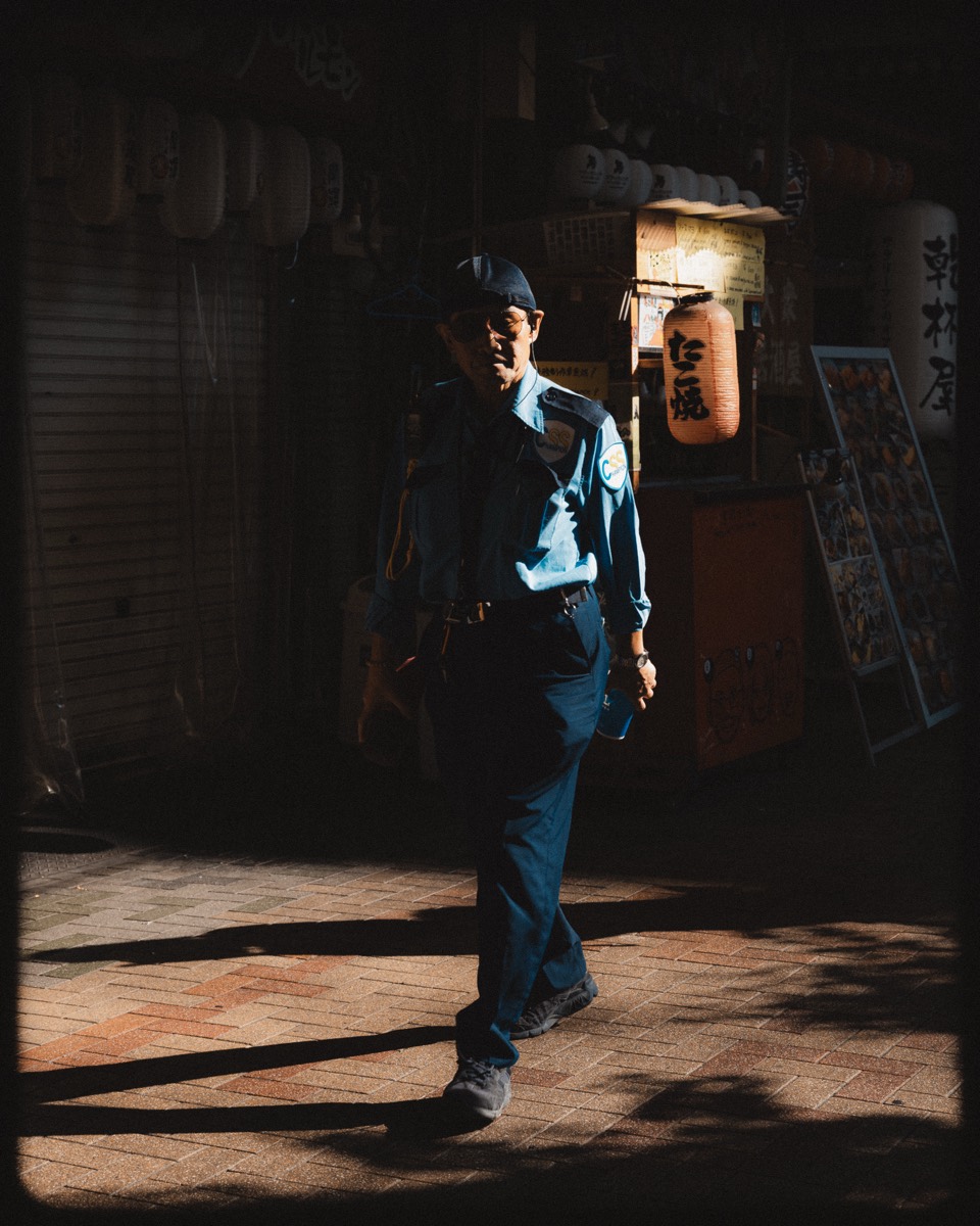A lone figure walks through a shaft of light past shuttered shopfronts and paper lanterns at night.